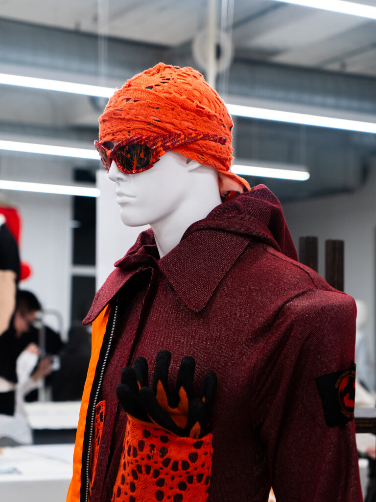 Mannequin with a textured burgundy jacket paired with an orange headscarf, matching sunglasses, and gloves tucked into a pocket.