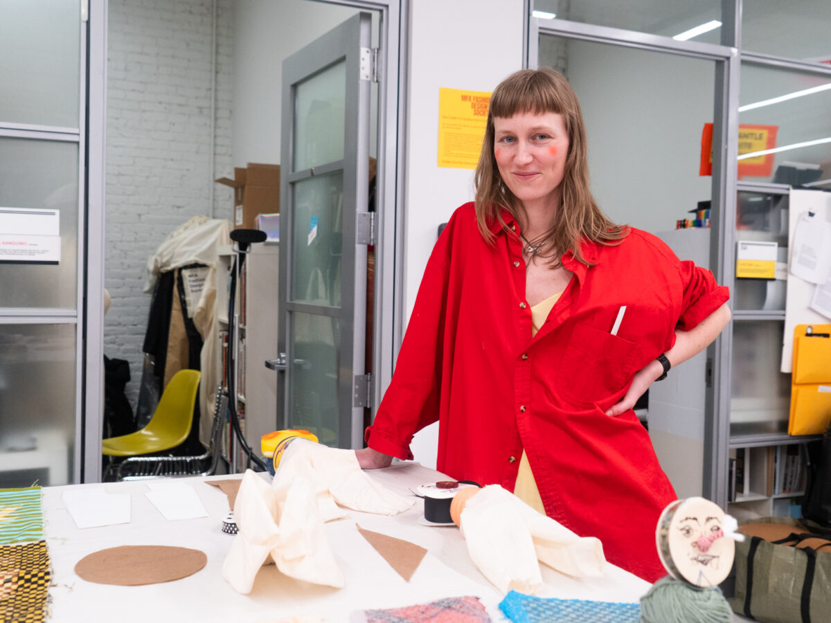  woman in a red shirt stands at a table covered with fabric samples, paper, and crafting materials.