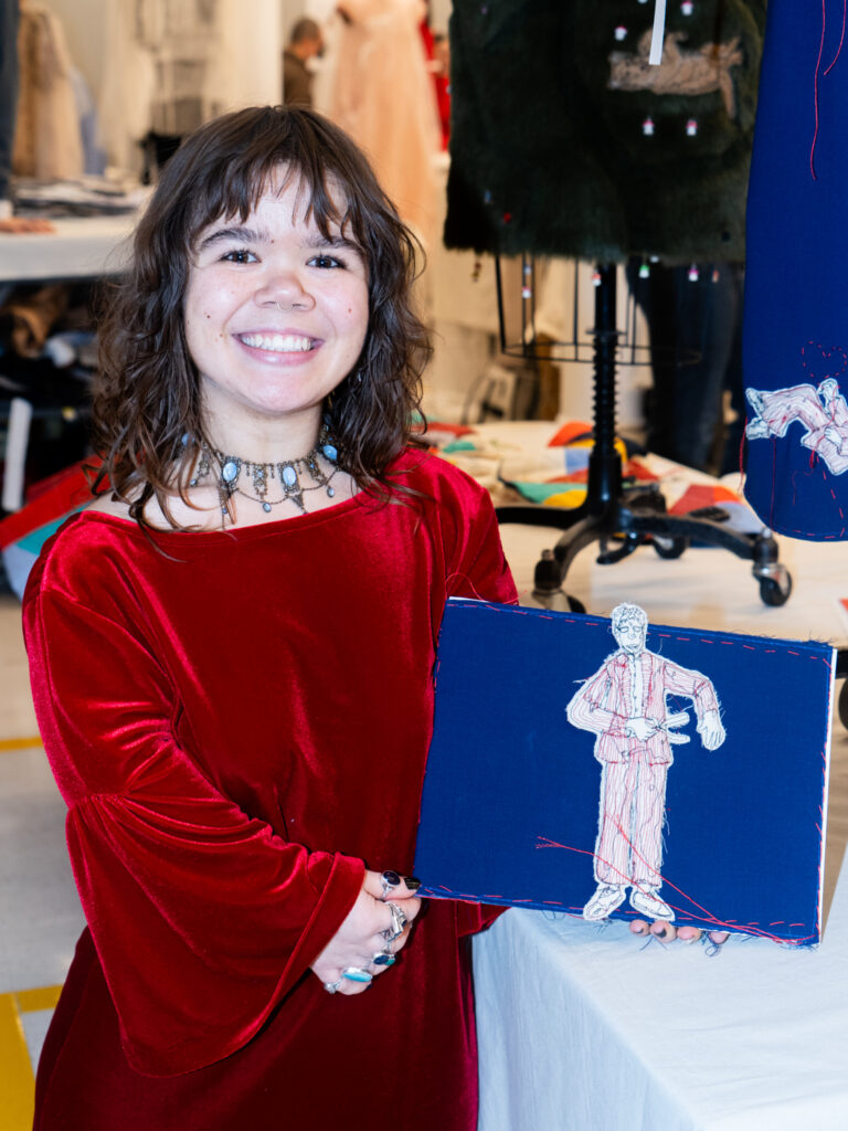 A woman in a red velvet dress holds up a stitched fabric artwork featuring an embroidered figure.