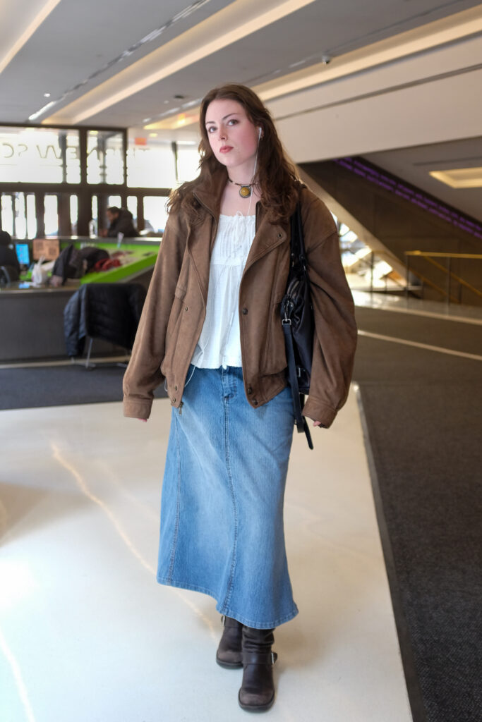 Student wears a brown jacket, a flowy white top, a maxi denim skirt, gray boots, a beaded necklace with a big multi-colored pendant and is carrying a black leather shoulder bag.