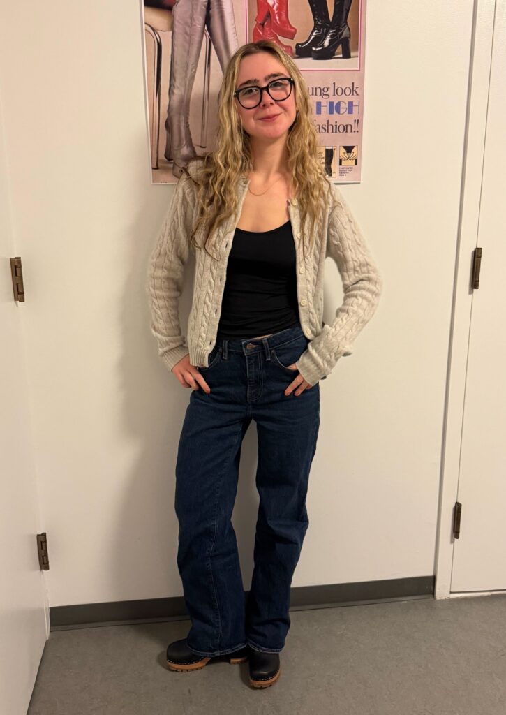 Student stands in front of white wall with pink poster of boots in the background. Student is wearing black glasses, a black tank top, a grey cardigan, dark wash blue jeans, and black boots. 