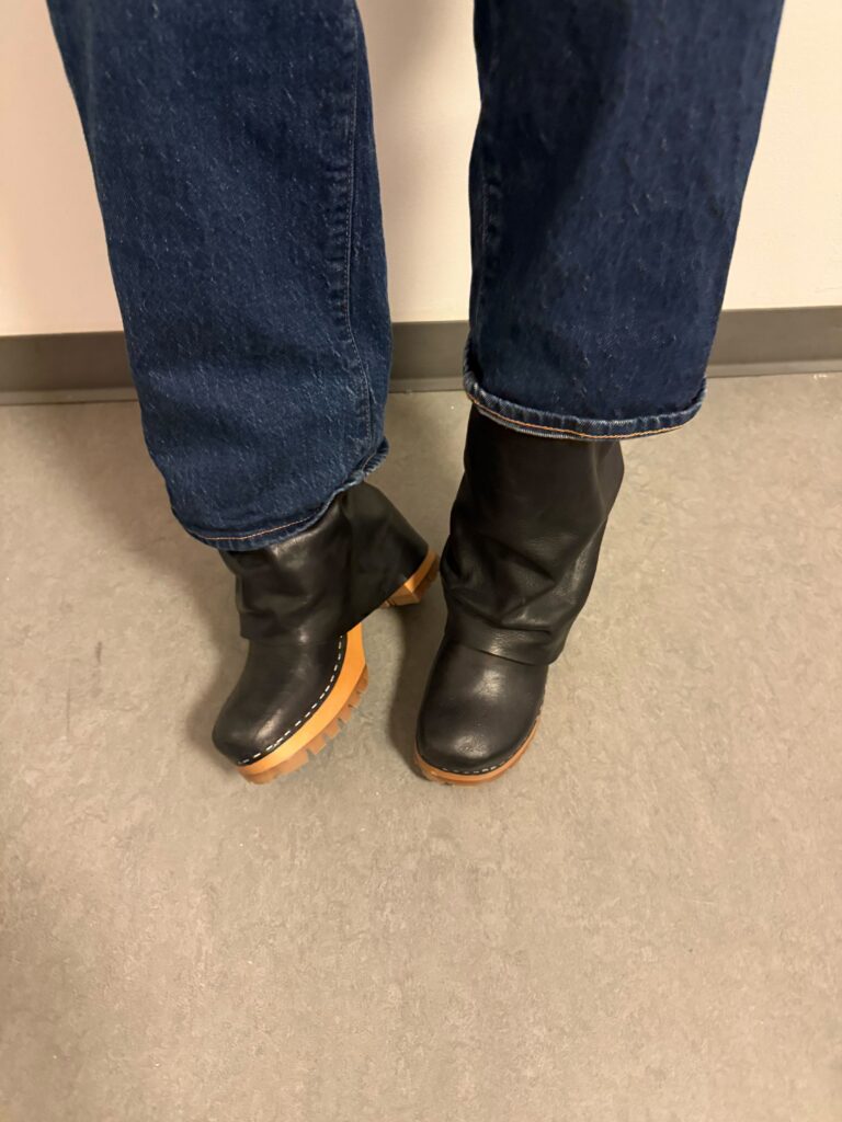 Close-up of a student standing on a grey floor with a white wall in the background wearing blue jeans and black, orange, and beige boots. 
