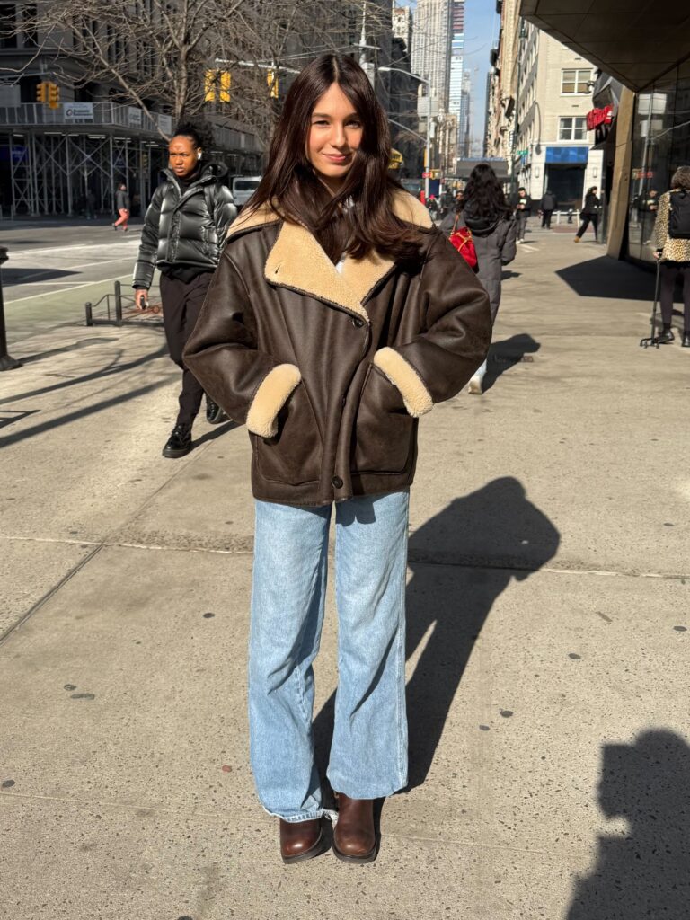 Student stands on Fifth Avenue wearing a brown coat, baggy blue jeans, and brown boots. 