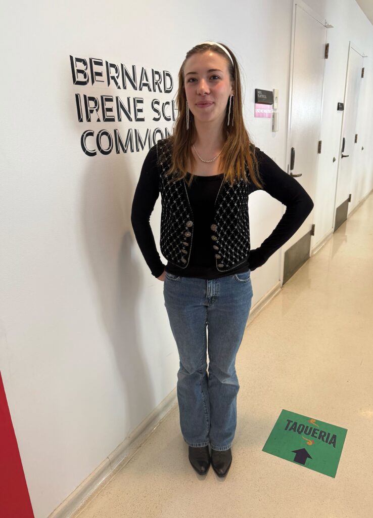 Student stands in front of a University Center cafeteria wall that is white with black text. The student is wearing a white headband, white dangling earrings, a black and white patterned vest, a black long sleeve top, bootcut blue jeans, and black cowboy boots. 