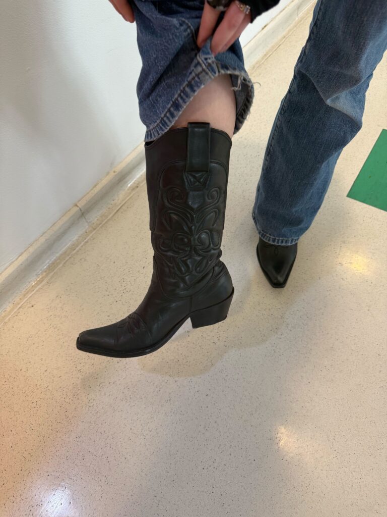 Close-up of a student standing on white floor with white background holding up their pant leg to show off a black detailed cowboy boot. 
