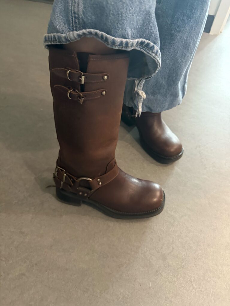 Close-up of a student standing on a grey floor wearing brown moto boots and blue jeans. 