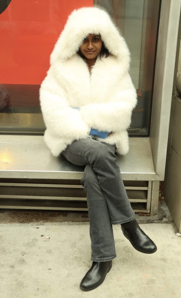 Student sits in front of the Sheila C. Johnson Design Center on 13th Street, wearing a white hooded fur coat, black jeans, and black boots. 