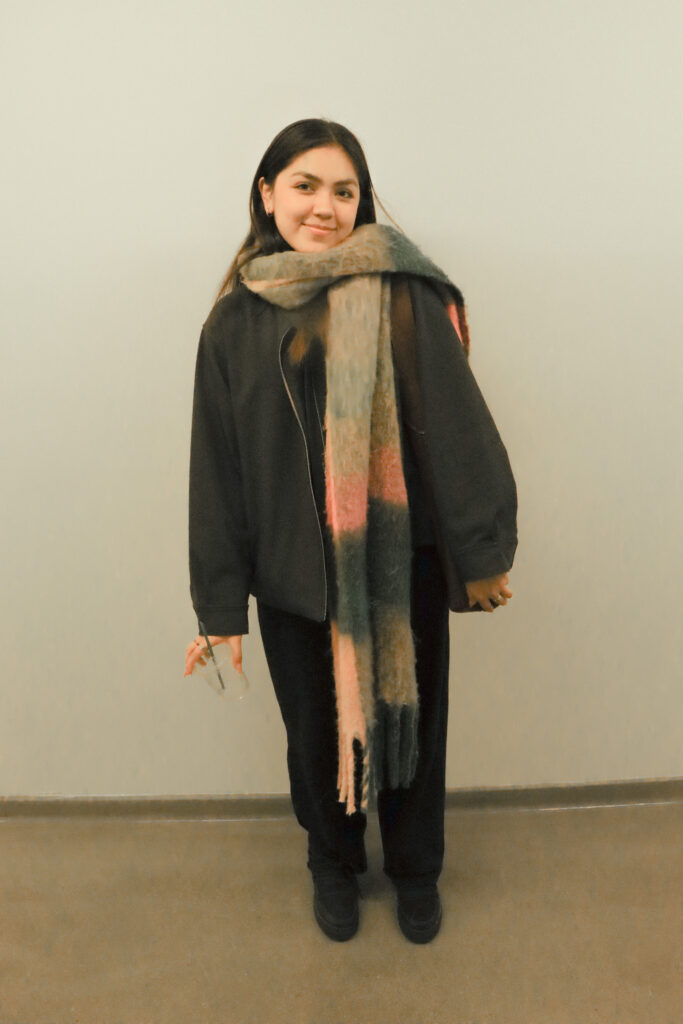 Student stands in front of a white wall in the University Center cafeteria wearing a black coat, multi-colored scarf, black pants, black boots, and a brown shoulder bag while holding a clear cup. 