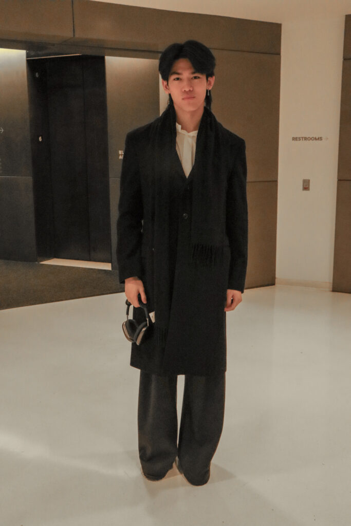 Student stands in front of the 6th floor University Center elevators wearing a black trench coat, a black scarf, a white button-up, black pants, and black shoes while holding a pair of headphones.