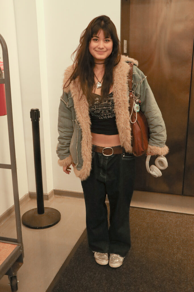 Student stands in front of the Kerrey Hall elevator wearing a silver heart necklace, a black tank top, a brown belt, black jeans, silver sneakers, a blue fur-lined coat, and holding a pair of white headphones. 