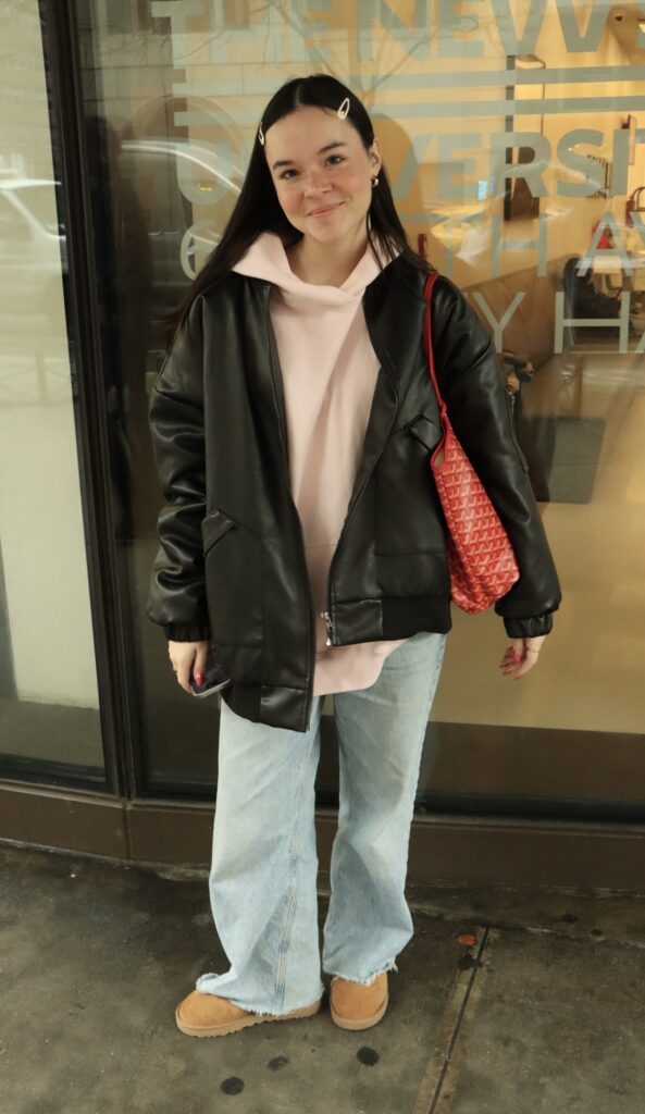 Student stands in front of the Kerrey Hall dorms wearing a pink hoodie, a black leather jacket, light-washed jeans, Uggs, and a red shoulder bag. 