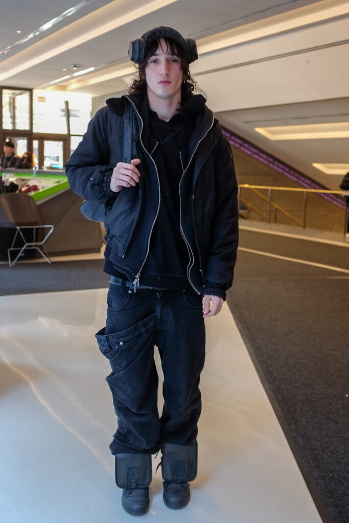 Student wears two black jackets, a black crewneck sweater, two pairs of black jeans with one unzipped and sagging downward, black shoes, a black pair of headphones and is carrying a denim tote backpack.