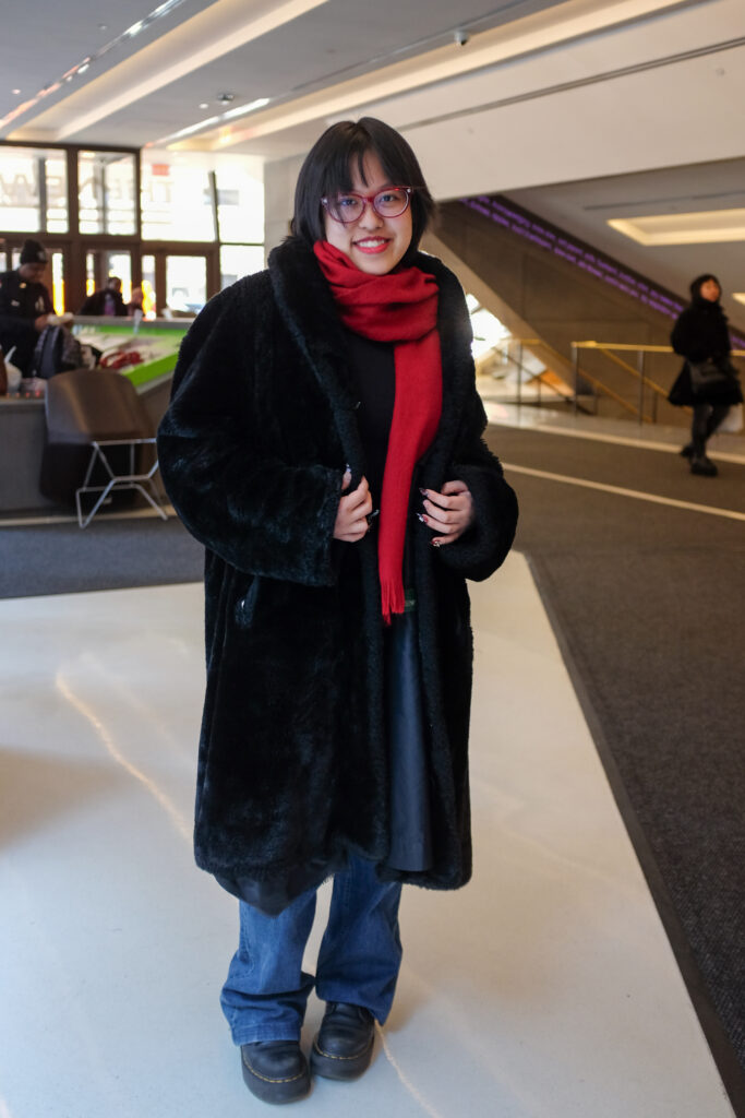 Student wears a black, faux fur trench coat, a chunky red scarf, baggy medium wash jeans, black Doc Martens, and a red pair of glasses.