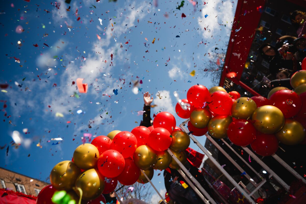 A hand reaches past red and gold balloons into the confetti-filled sky. 