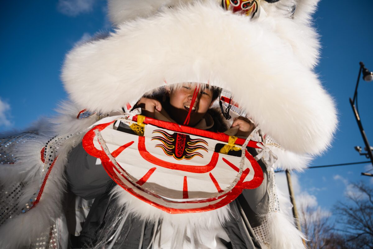 A young performer peers out of the mouth of a white, furry dragon puppet against the blue sky. 