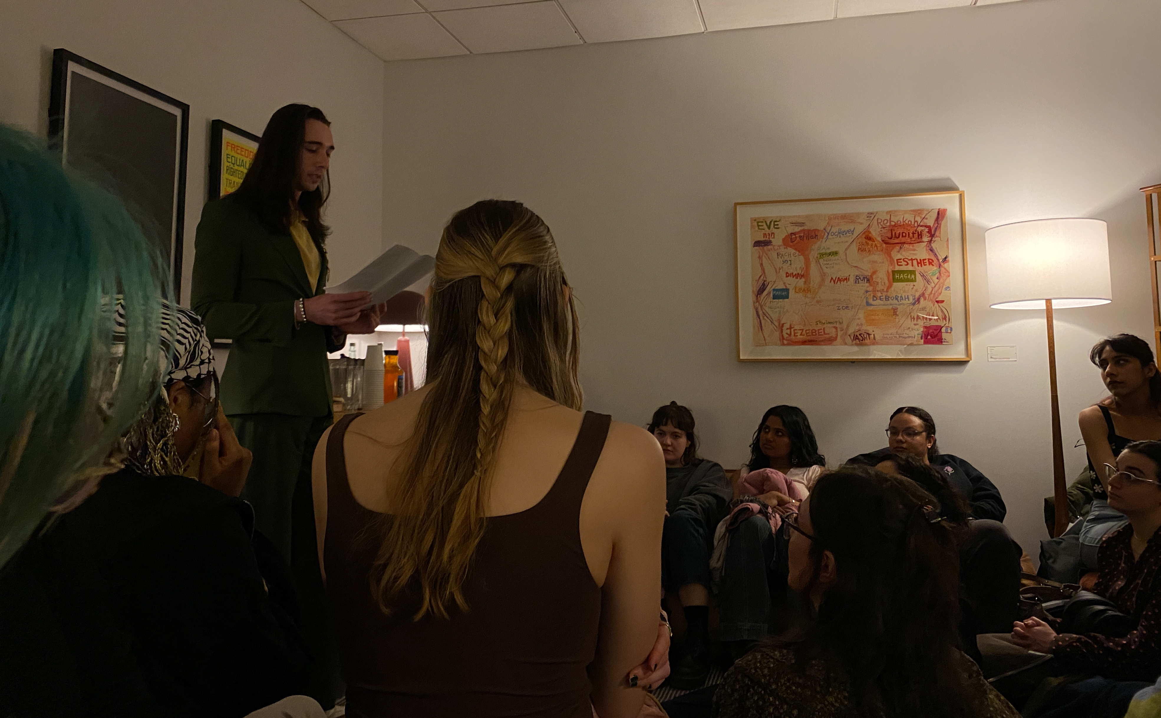 A person, standing, holding a piece of paper, in a dimly lit room, in front of a seated audience.