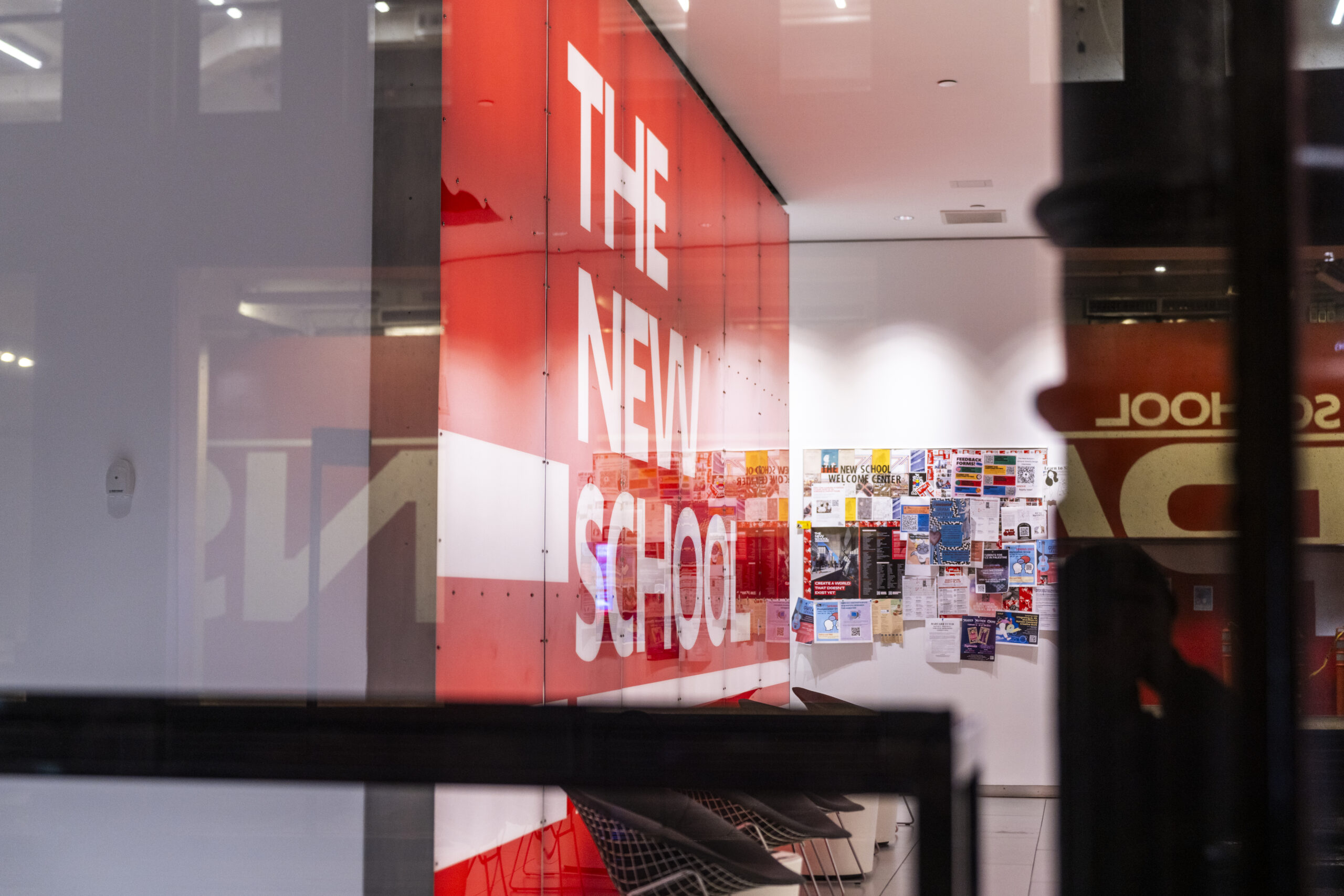 Side view through a window of The New School logo in white text on a red wall. Several posters hang on a far wall in the background. The Parsons School of Design logo is reflected on the window