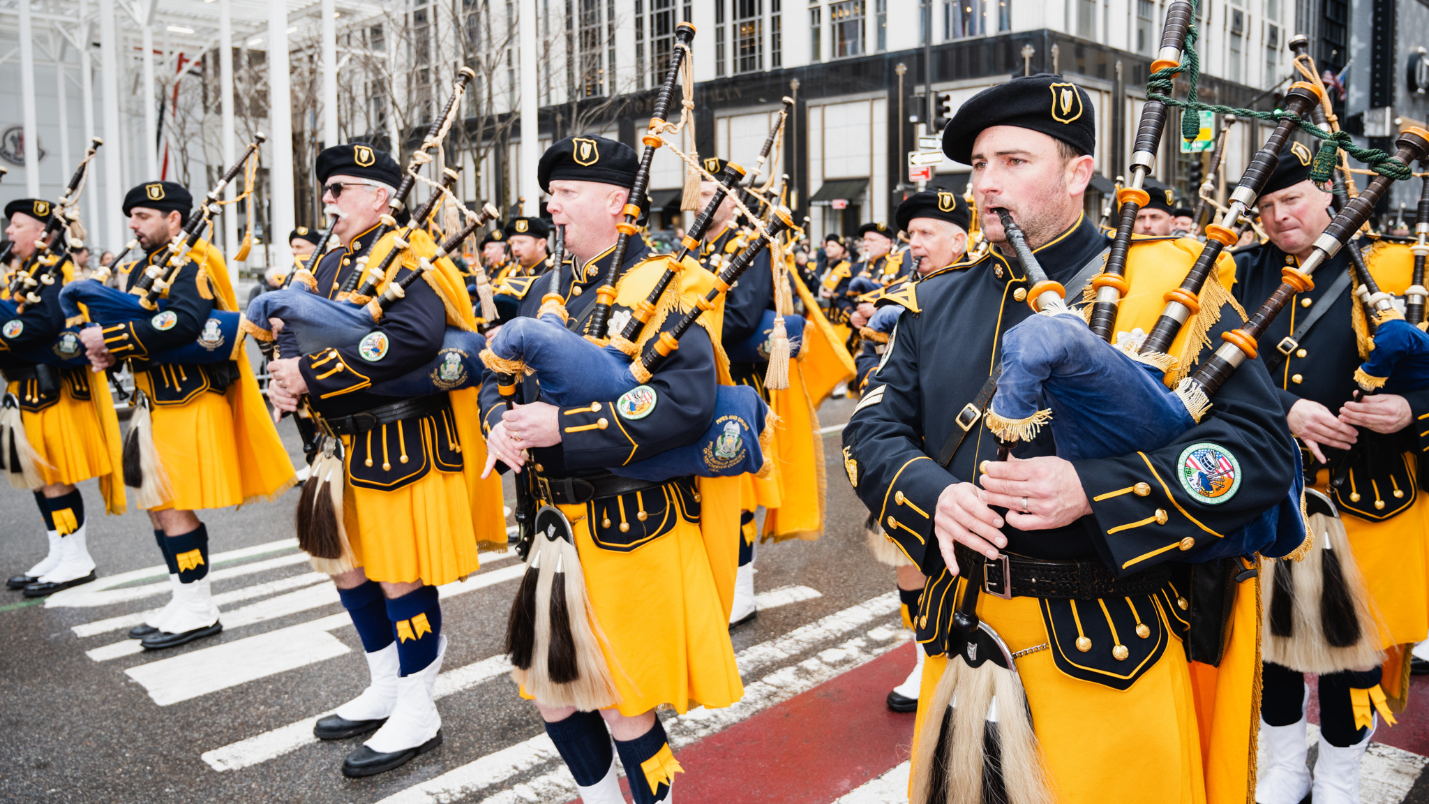 Rows of bagpipe performers dressed in yellow and navy kilts march along the parade route while playing their bagpipes.